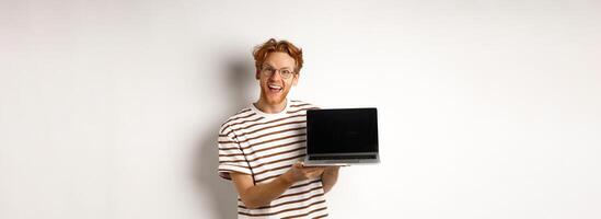 Funny redhead man in glasses showing laptop screen advertisement and smiling. Guy with red hair demonstrates promo or banner on display, white background photo
