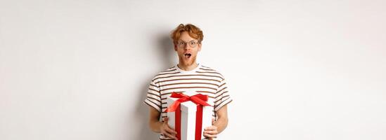 Valentines day and holidays concept. Surprised redhead boyfriend in glasses looking grateful at camera, receive big present in box, standing over white background photo