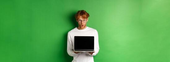 Impressed redhead nerdy guy in glasses showing blank laptop screen and looking at display amazed, standing over green background photo