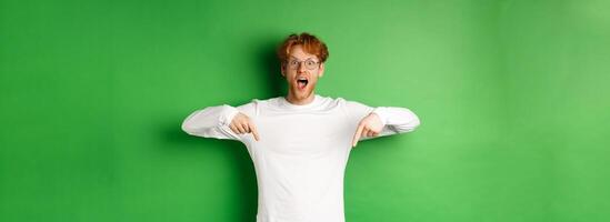 Surprised and excited young redhead man scream of joy and amazement, checking out awesome promo, pointing fingers down, standing over green background photo