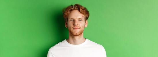Close-up of young man with red messy hair and beard looking at camera, standing over green background photo