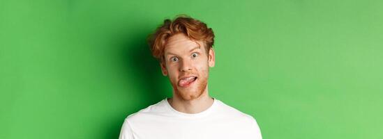 Emotions and fashion concept. Close up of funny redhead man showing silly faces, sticking tongue and staring at camera, standing over green background photo