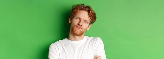 Emotions and fashion concept. Close-up of skeptical young man with red hair and beard pucker lips, looking at camera as waiting for something, standing over green background photo