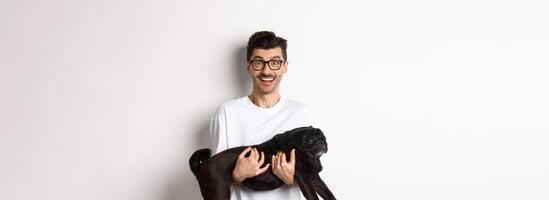Happy young man in glasses holding black pug pet and staring at camera amused, smiling cheerful, standing over white background photo