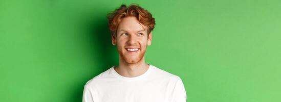 Headshot of happy redhead man with beard wearing white long sleeve, looking left at copy space and smiling, standing over green background photo