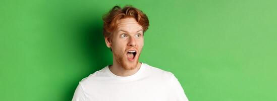 Close up of surprised and impressed redhead man checking out promotion offer, looking left with dropped jaw, standing over green background photo