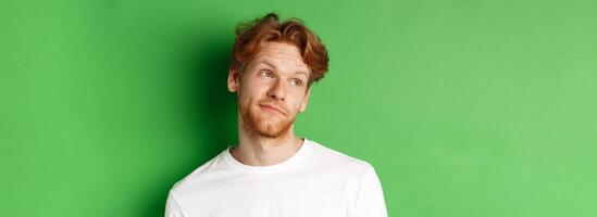 Smiling redhead man with messy haircut and beard tilt head, looking left with pleased face, standing over green background photo