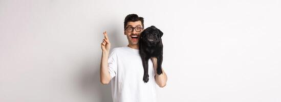Hopeful smiling dog owner making a wish, holding cute black pug on shoulder and cross fingers for good luck, white background photo