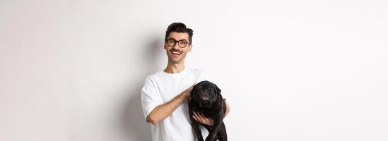 Happy hipster guy in glasses pet dog and smiling. Cute black pug enjoy spending time with owner, looking satisfied, standing over white background photo