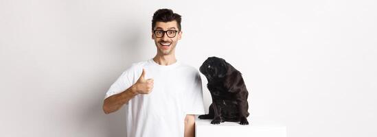 Cheerful handsome guy standing near cute black pug and showing thumb-up. Pet owner approve and recommend product for dogs, white background photo