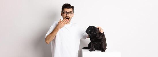 Image of surprised and happy dog owner pet his dog and staring at camera with rejoice, standing with pug over white background photo