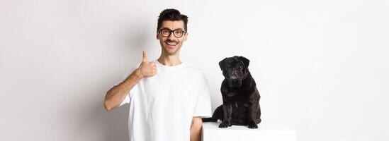 Image of dog owner and cute black pug looking at camera, man showing thumb-up in approval, recommending something, standing over white background photo