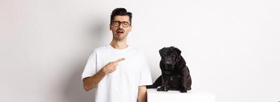 Upset crying man pointing at cute black pug and sobbing, complaining on his pet, standing sad against white background photo
