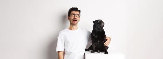Whining man looking sad while pug staring intrigued at upper left corner, standing against white background photo