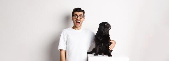 Image of amazed man staring at camera while his small dog looking left at copy space, standing over white background photo