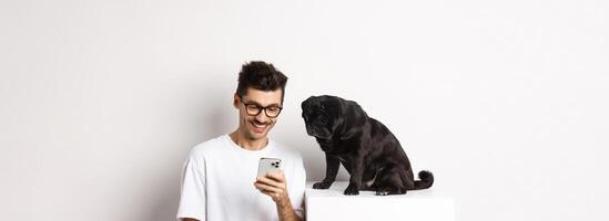 Smiling young man using smartphone and sitting near dog. Pug owner checking photos on mobile phone, white background