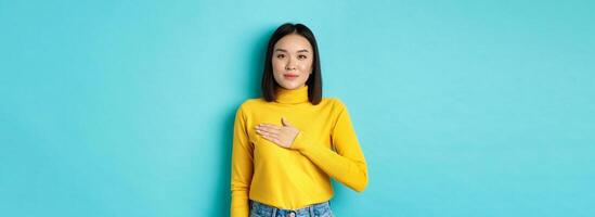 Image of proud smiling asian woman holding hand on heart, showing respect to national anthem, standing over blue background photo