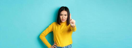 Confident and serious woman tell no, showing extended finger to stop and prohibit something bad, frowning and looking at camera self-assured, standing over blue background photo