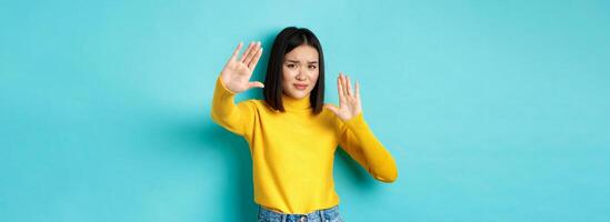 Image of woman feeling uncomfortable in front of camera, cover face with hands and frowning, begging to stop, standing against blue background photo