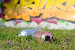 Several used spray cans with paint and caps for spraying paint under pressure on grass near the painted wall in colored graffiti photo