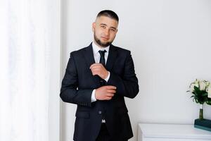 A man in a suit and tie is standing in front of a window, adjusting his tie. Concept of formality and professionalism, as the man is dressed in a suit and tie photo