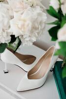 A pair of white high heels are displayed on a table next to a vase of flowers. The shoes are pointed and elegant, and the flowers add a touch of freshness and color to the scene photo
