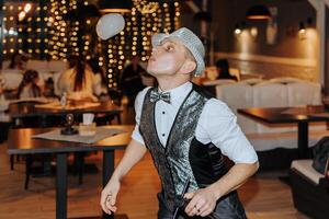 A man in a black vest and hat is juggling a ball. The scene takes place in a restaurant with tables and chairs photo