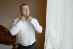 A man in a white shirt is getting dressed in front of a window. Scene is one of preparation and focus, as the man is carefully adjusting his collar and shirt photo