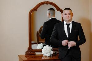A man in a suit stands in front of a mirror, adjusting his tie. Concept of formality and elegance, as the man is dressed in a suit and tie, and the mirror reflects his appearance photo