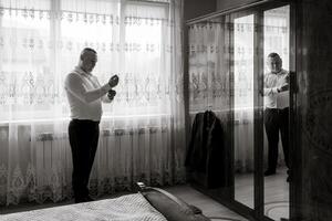 A man is getting ready for a formal event, putting on his suit and tie. He is standing in front of a mirror, adjusting his tie and looking at himself. The room is decorated with curtains and a bed photo