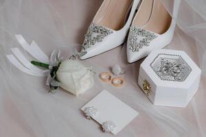 A pair of white shoes with a rose on top of them. A white box with a ring inside photo