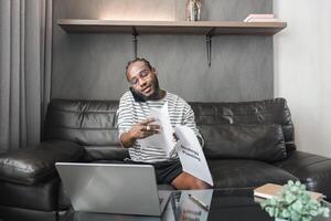 Young African American Man Working from Home on Couch with Laptop and Documents, Talking on Phone photo