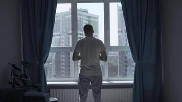 Rear view of a pensive man standing at home by the window and looking at the city. Media. Man in home clothes spending time in reflection in his bedroom. photo