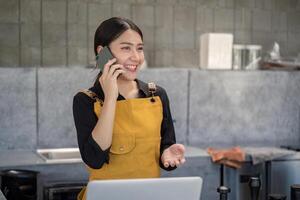 Young Barista in Yellow Apron Smiling While Talking on Smartphone in Modern Kitchen photo
