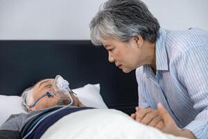 Senior fatal dying male patient with oxygen mask is getting consoling support from his grieving wife while suffering from final stage cancer at home with palliative medical treatment in his final hour photo