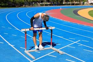 Senior Asian athlete is practicing hurdling running in the professional blue track for professional exercise, sport training workout and healthy for longevity concept photo