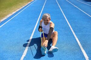 Senior Asian athlete using stretching rubber band warm up for running in the professional blue track for professional exercise, sport training workout and healthy for longevity photo