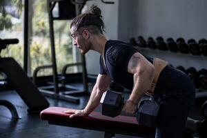 Caucasian man with strong and muscular body training on bent over row dumbbell with bench to build back and wing muscle inside the gym with dark tone style photo