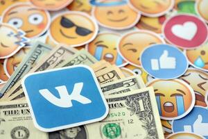 KYIV, UKRAINE - APRIL 20, 2024 VK logo and many usd money bills. Symbol of famous social network platform and dollar currency banknotes on lot of emoji reactions photo
