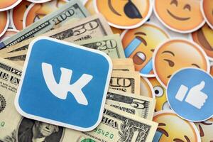 KYIV, UKRAINE - APRIL 20, 2024 VK logo and many usd money bills. Symbol of famous social network platform and dollar currency banknotes on lot of emoji reactions photo