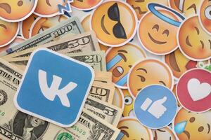 KYIV, UKRAINE - APRIL 20, 2024 VK logo and many usd money bills. Symbol of famous social network platform and dollar currency banknotes on lot of emoji reactions photo