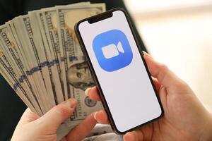 KYIV, UKRAINE - APRIL 1, 2024 Zoom icon on smartphone screen and money in female hand photo