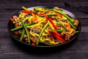 Appetizing stir fried noodles with garlic sprouts, mushrooms, red pepper and chicken meat on a black rustic background of a wooden table. A popular Asian dish photo