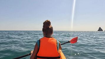A young girl in an orange life vest rows a kayak on the open water. A distant island is visible on the horizon. The sky is blue and sunny. video
