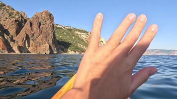 A hand reaches out from a yellow kayak towards the rocky cliffs lining the coast, with blue water in the foreground. video