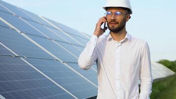 Confident engineer wearing safety hard hat, holding mobile phone, making a call and walking. Engineer, worker, or architect working with Solar panel concept video