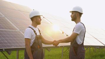 Two workers engineers in helmet and overalls shaking hands after discussion on background of photovoltaic solar panels video