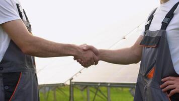 Two engineers shaking hands after discussion on background of photovoltaic solar panels, close up video