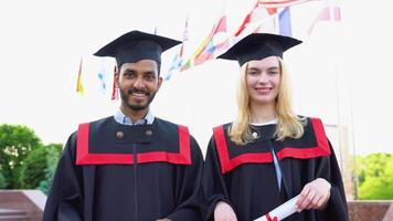 Portrait of happy multiracial graduates. Two friends in graduation caps and gowns standing outside university building, holding diploma scrolls, and smiling video