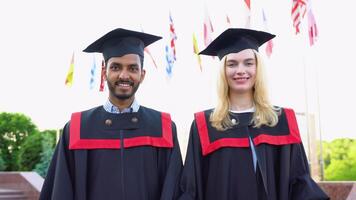 Portrait of happy multiracial graduates. Two friends in graduation caps and gowns standing outside university building, holding diploma scrolls, and smiling video